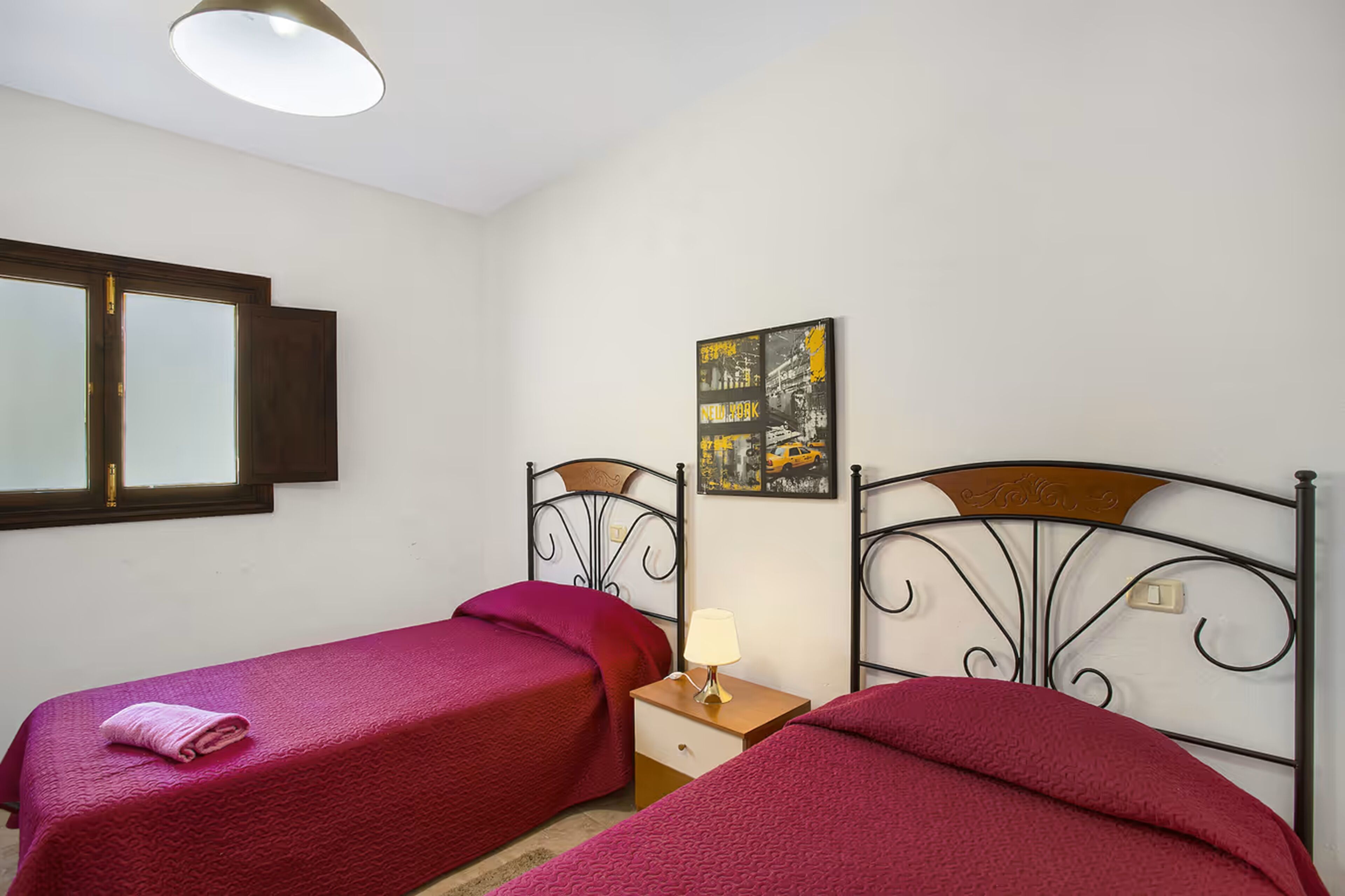 A bedroom with two single beds, a nightstand with a lamp, a window with blinds, and a wall-mounted artwork.