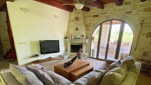 A cozy living room with a stone fireplace, a wooden ceiling, and a large window. - Traditional stone house for rent in Crete in a quiet location (Vamos)
