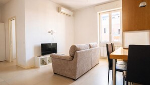 A modern living room with a beige sofa, a flat-screen TV, a dining table with chairs, and a large window with a view of buildings.