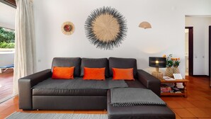 Un salon moderne avec un canapé foncé, des coussins orange, un élément décoratif rond fixé au mur, une petite table avec une lampe, et une vue sur l’extérieur.