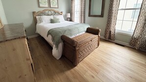 Une chambre avec un plancher en bois, un lit recouvert d’une couverture verte, un banc en osier, une commode et une fenêtre avec des rideaux.
