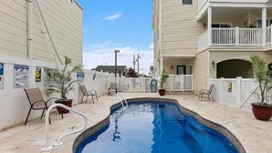 Une piscine dotée d’une terrasse blanche, entourée de plantes en pot et de chaises, avec un bâtiment d’un côté et une clôture de l’autre.