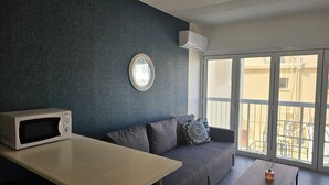 A compact living space with a kitchenette, a grey sofa with patterned cushions, a round mirror, and a large sliding glass door to a balcony.