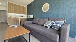 A modern living room with a grey sofa, a wooden coffee table, and a kitchen area with wooden cabinets and a white countertop.
