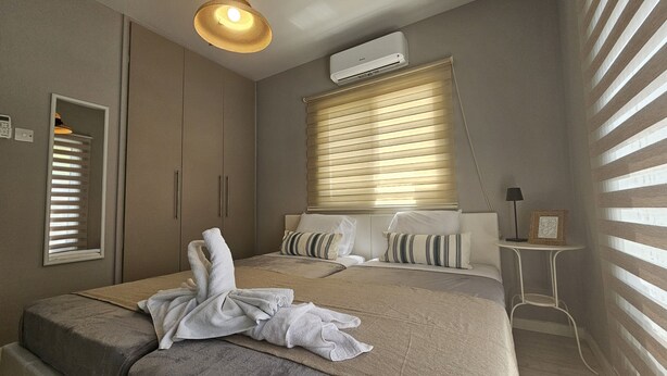 A bedroom with a bed, a bedside table, a lamp, a mirror, and a window with blinds.