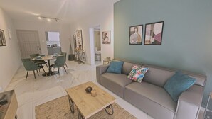 A modern living room with a grey sofa, green chairs, a wooden coffee table, and a kitchen in the background.