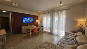 A living room with a TV on the wall, a dining table with chairs, a sofa, and a lamp. - 62 APH - Stay Aphrodite Hills Apartment (Kouklia)
