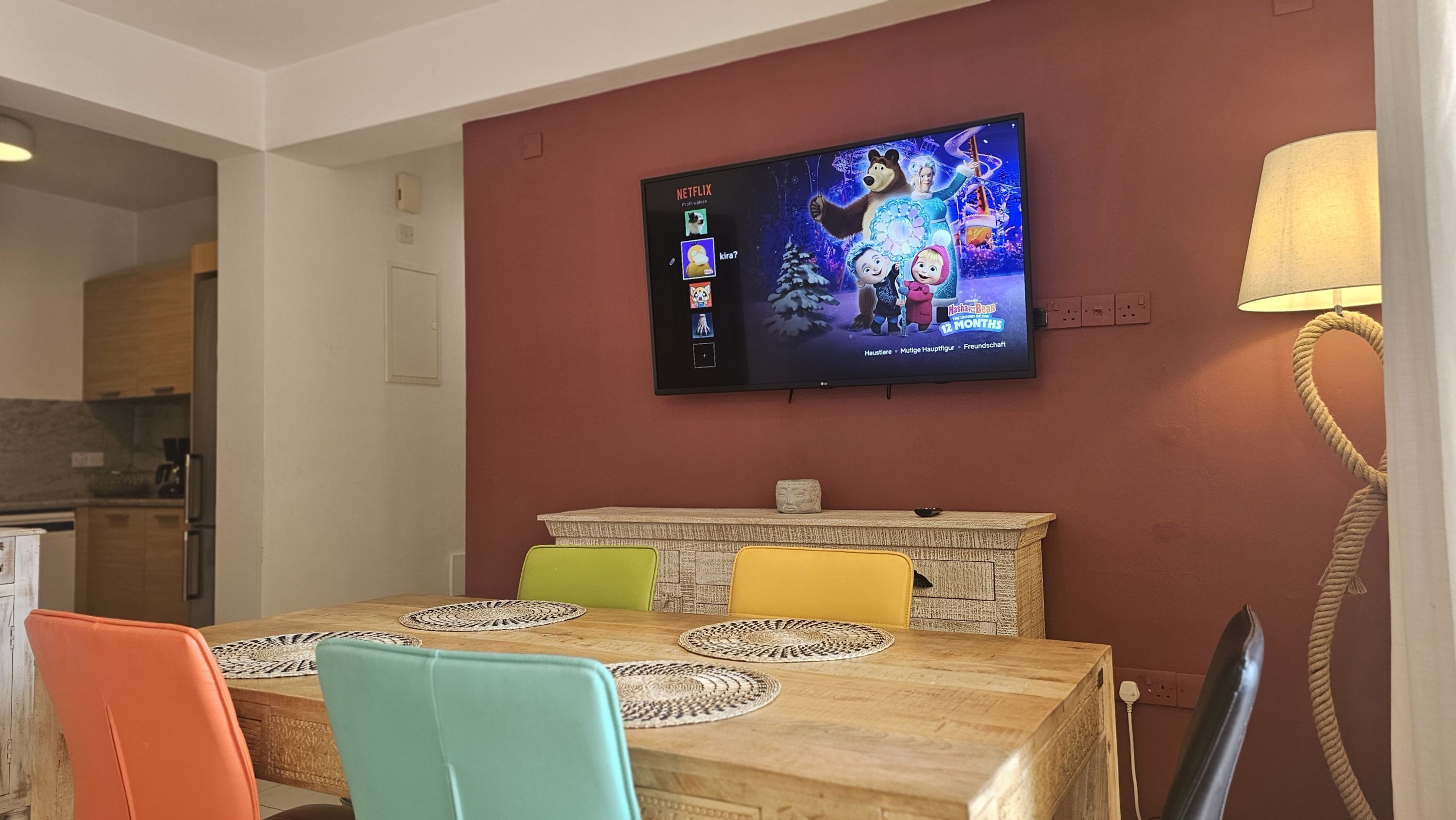 Een eetruimte met een houten tafel, kleurrijke stoelen, een aan de muur bevestigde televisie en een lamp.
