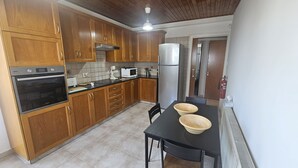 Cocina con armarios de madera, encimera negra, microondas, estufa y refrigerador. Hay una mesa con dos tazones y una silla.