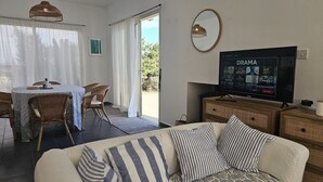 Ein Wohnzimmer mit Fernseher, der einen Streaming-Dienst anzeigt, ein Essbereich mit Korbmöbeln und Blick durch eine Glastür.