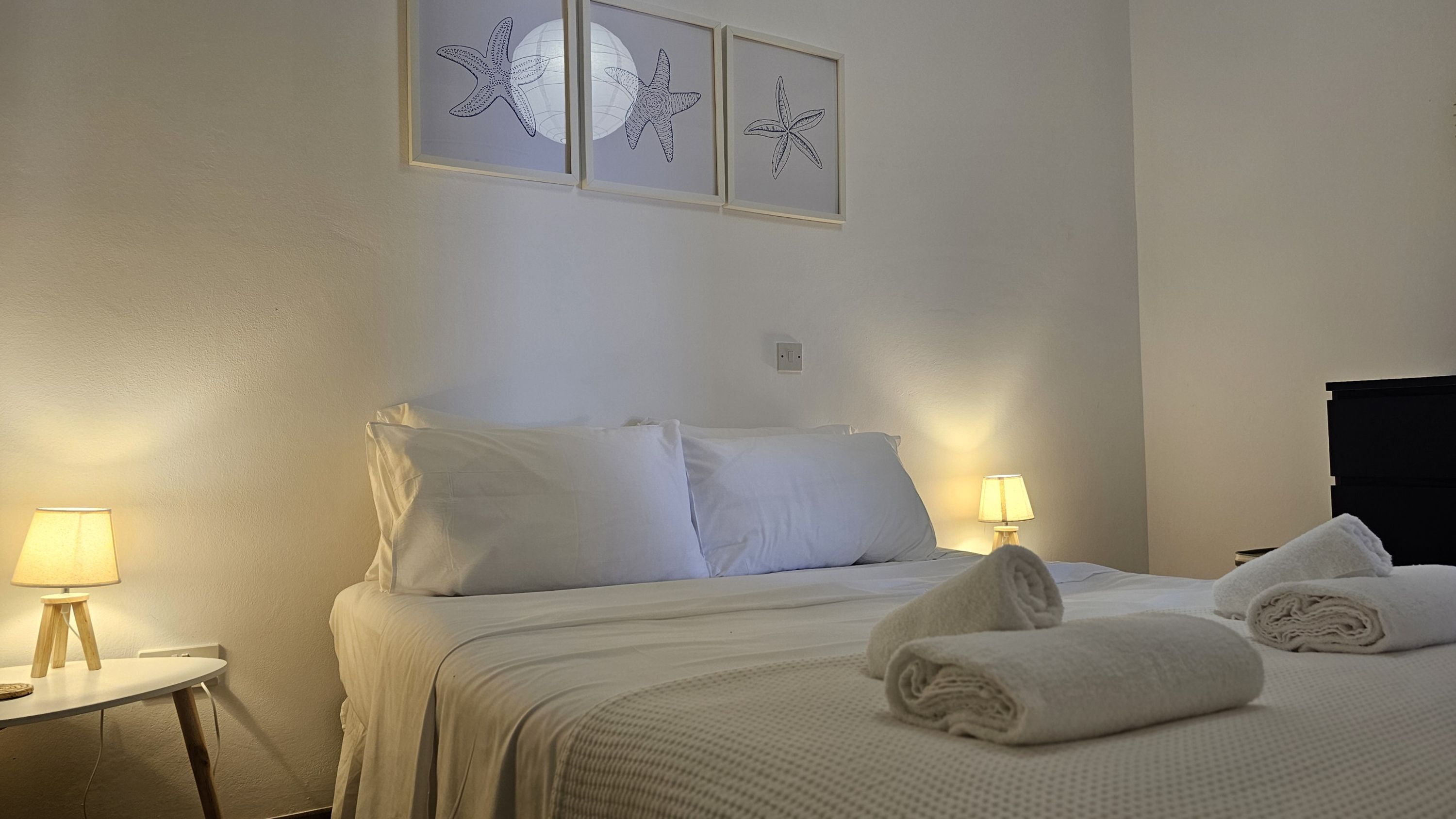 A neatly made bed with white linens, two bedside lamps, and three framed starfish art pieces on the wall.