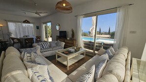 Ein geräumiges Wohnzimmer mit einer Sofagarnitur, einem Couchtisch und einem Fernseher. Es gibt einen Essbereich mit Tisch und Stühlen sowie einen Balkon mit Blick auf den Pool.