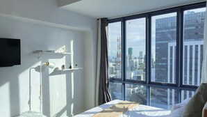 A modern hotel room with a large window offering a city view, a bed with a white and brown blanket, a bedside table, and a mounted TV. - Cozy studio with Pool and fitness room in enjoyable Cebu City (Cebu City)
