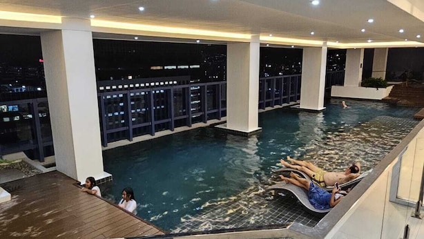 A modern indoor swimming pool with a wooden deck area, surrounded by glass railings and a cityscape view. - Cozy studio with Pool and fitness room in enjoyable Cebu City (Cebu City)