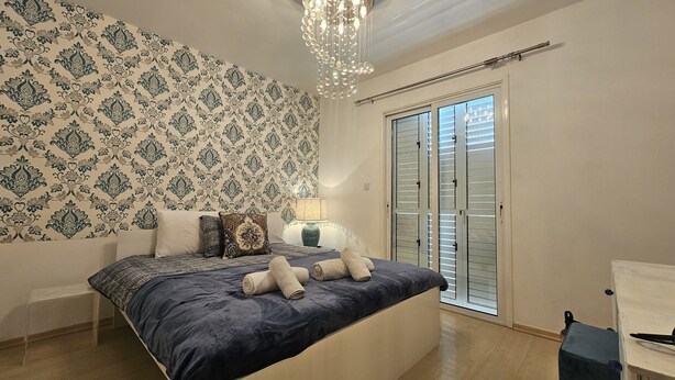 A bedroom with a patterned wallpaper, a bed with rolled towels, a bedside lamp, and a sliding glass door to the right. - 27 NIC - Stay Alex s House (Strovolos)