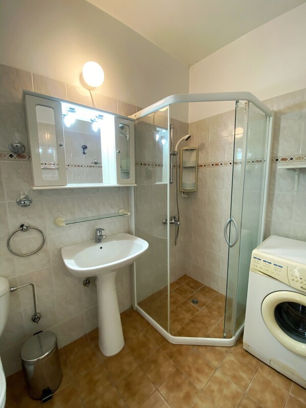 A bathroom with a glass-enclosed shower, a pedestal sink, a toilet, a washing machine, and a towel rack.