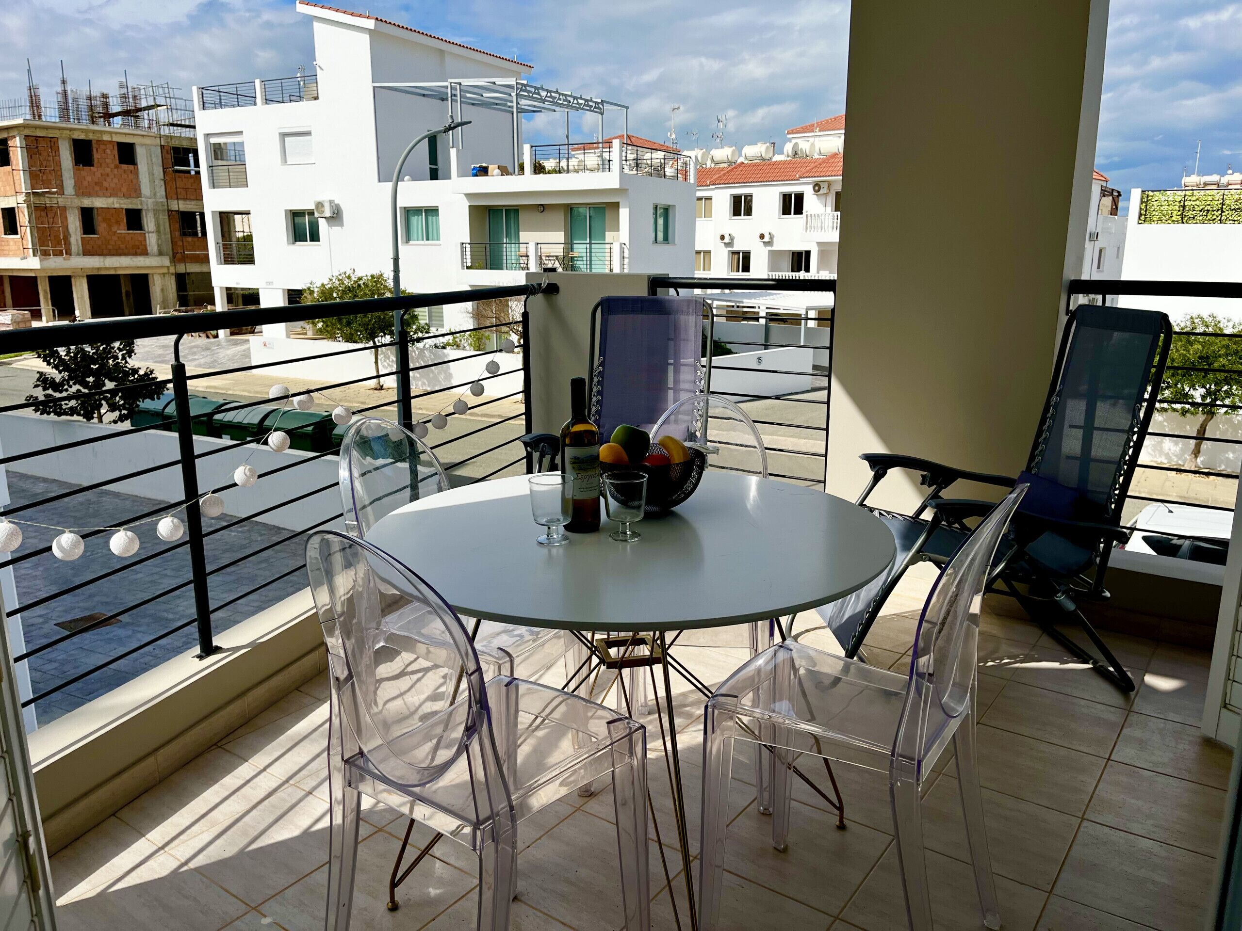 A balcony with a round table and clear plastic chairs, overlooking a street with buildings under construction.