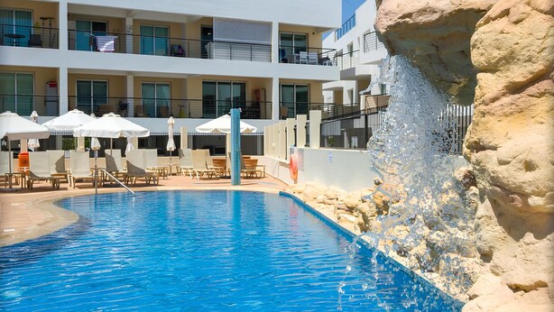 Une piscine avec des chaises longues et des parasols, un bâtiment doté de balcons et une formation rocheuse.