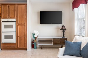 Een moderne keuken met houten kastjes en een inbouwoven, een flatscreen televisie aan de muur, een witte console tafel en een witte bank met blauwe kussens.