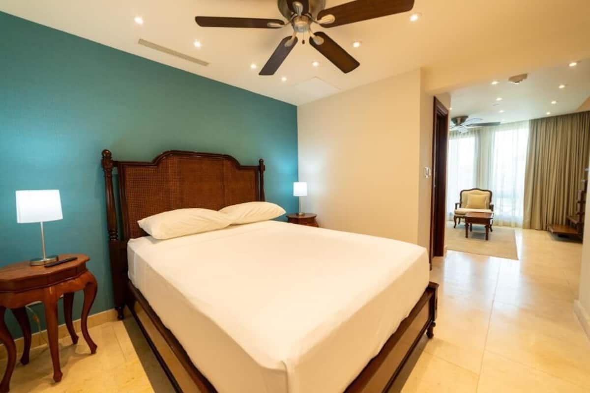 A hotel room with a large bed, wooden headboard, two bedside tables with lamps, a ceiling fan, and a view of a balcony.