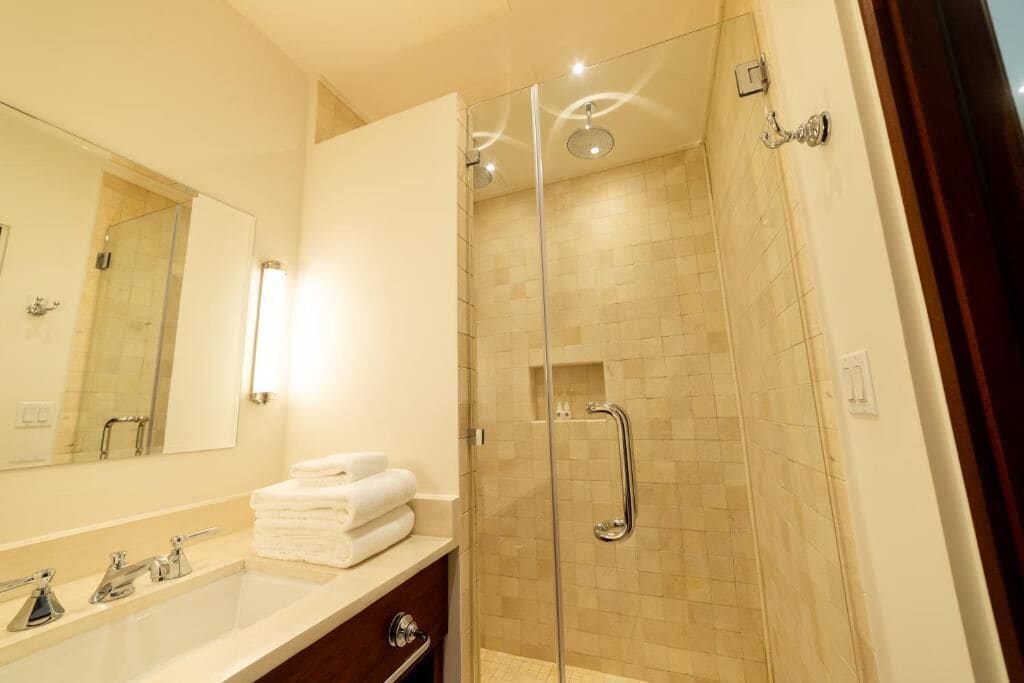 A modern bathroom with a glass-enclosed shower, a large mirror, and a stack of neatly folded towels on the vanity.