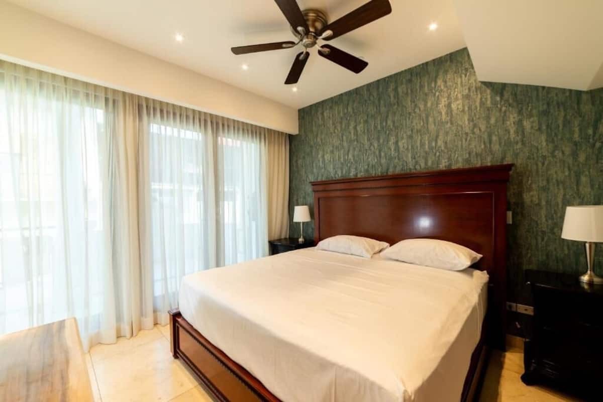 A hotel room with a large bed, a ceiling fan, a nightstand with a lamp, and a view of the outdoors through sheer curtains.
