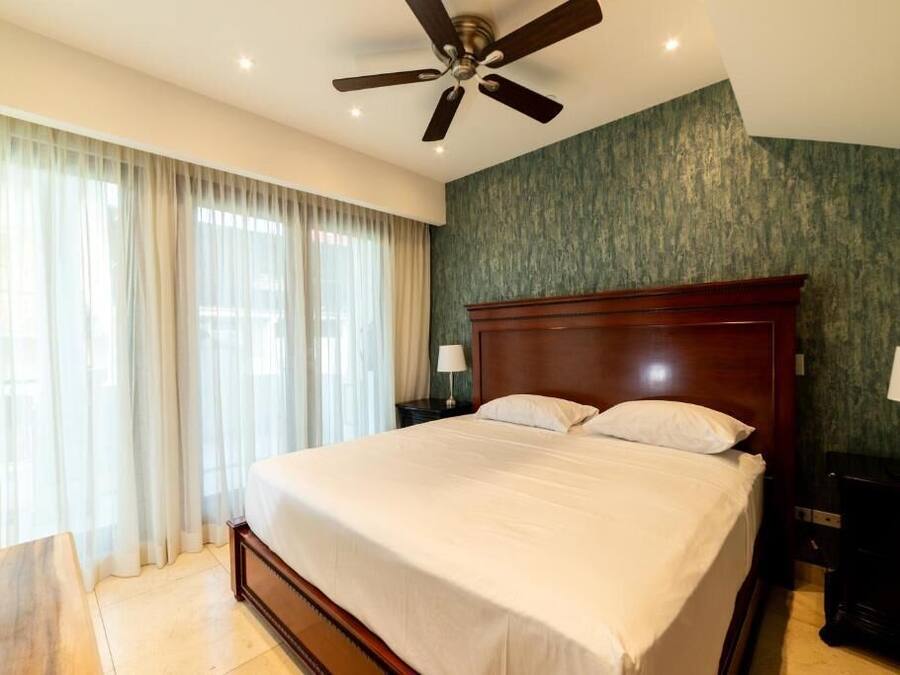 A hotel room with a large bed, a ceiling fan, a nightstand with a lamp, and a view of the outdoors through sheer curtains.