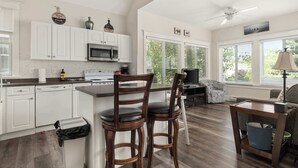 A kitchen with white cabinets, a microwave, and a stove. There are two wooden chairs with black cushions. A ceiling fan is visible, and a window with blinds is on the wall.