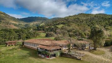 Um edifício grande e rústico com telhado de telhas, rodeado por vegetação exuberante e colinas.