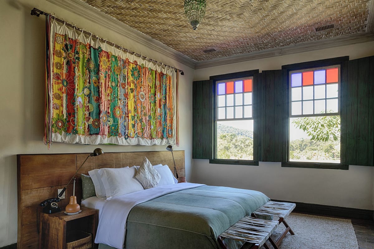 A bedroom with a wooden headboard, a bed with a green bedspread, a nightstand with a lamp, and a colorful tapestry.