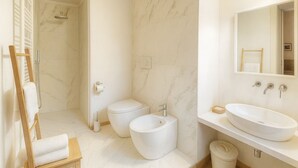 Een moderne badkamer met een marmeren vloer, een houten stoel, twee toiletten, een grote wasbak en een douche.