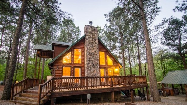Eine Blockhütte mit einem Steinkamin, einer Holzterrasse und Blick auf den umliegenden Wald.