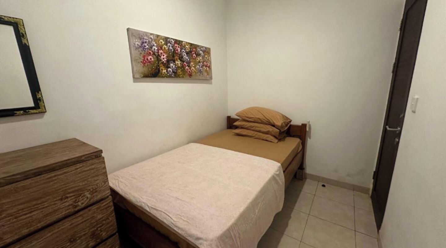 Ein kleines, ordentlich eingerichtetes Schlafzimmer mit einem Bett, einer Holzkommode und einem an der Wand hängenden Bild.