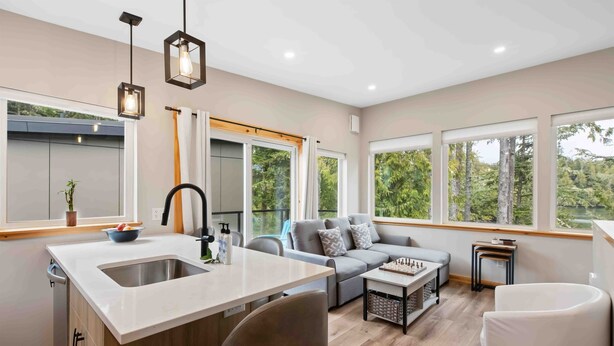 A modern kitchen with a central island, a dining area with a sofa, and large windows offering a view of the outdoors. - SS#5-Shortboarder-Spring Cove View, Sauna, Hikes! (Ucluelet)