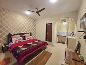 A bedroom with a bed, a bedside table, a desk, a chair, a ceiling fan, a wardrobe, and a bathroom visible through an archway. - Himalayan Horizon and Cafe (Dharamshala)