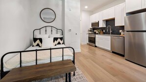 Une chambre à coucher moderne, avec un lit, une table de chevet et un grand miroir. À côté se trouve une cuisine avec des armoires blanches, des appareils en acier inoxydable et un plancher en bois.