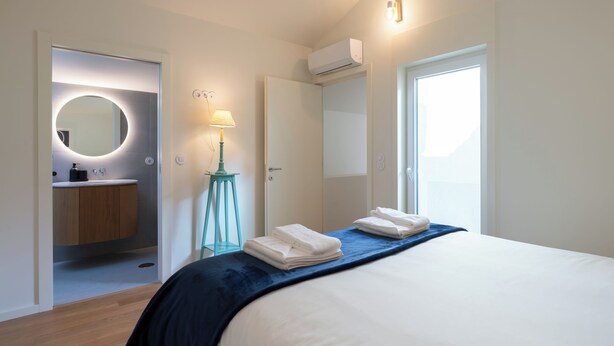 Ein modernes Hotelzimmer mit Bett, einem Badezimmer mit rundem Spiegel, einer Stehlampe und einem Fenster, das für natürliches Licht sorgt.