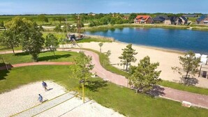 A sandy beach area with a volleyball net, trees, and a lake in the background. - Appartement im Wangerland Resort (Hohenkirchen)