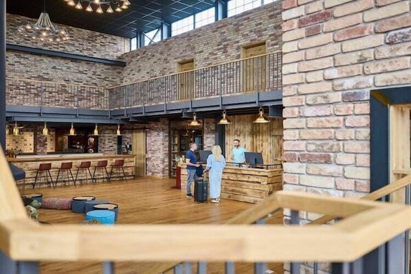 A spacious lobby with a high ceiling, exposed brick walls, and a wooden reception desk. There are multiple seating areas with chairs and a bar counter.
