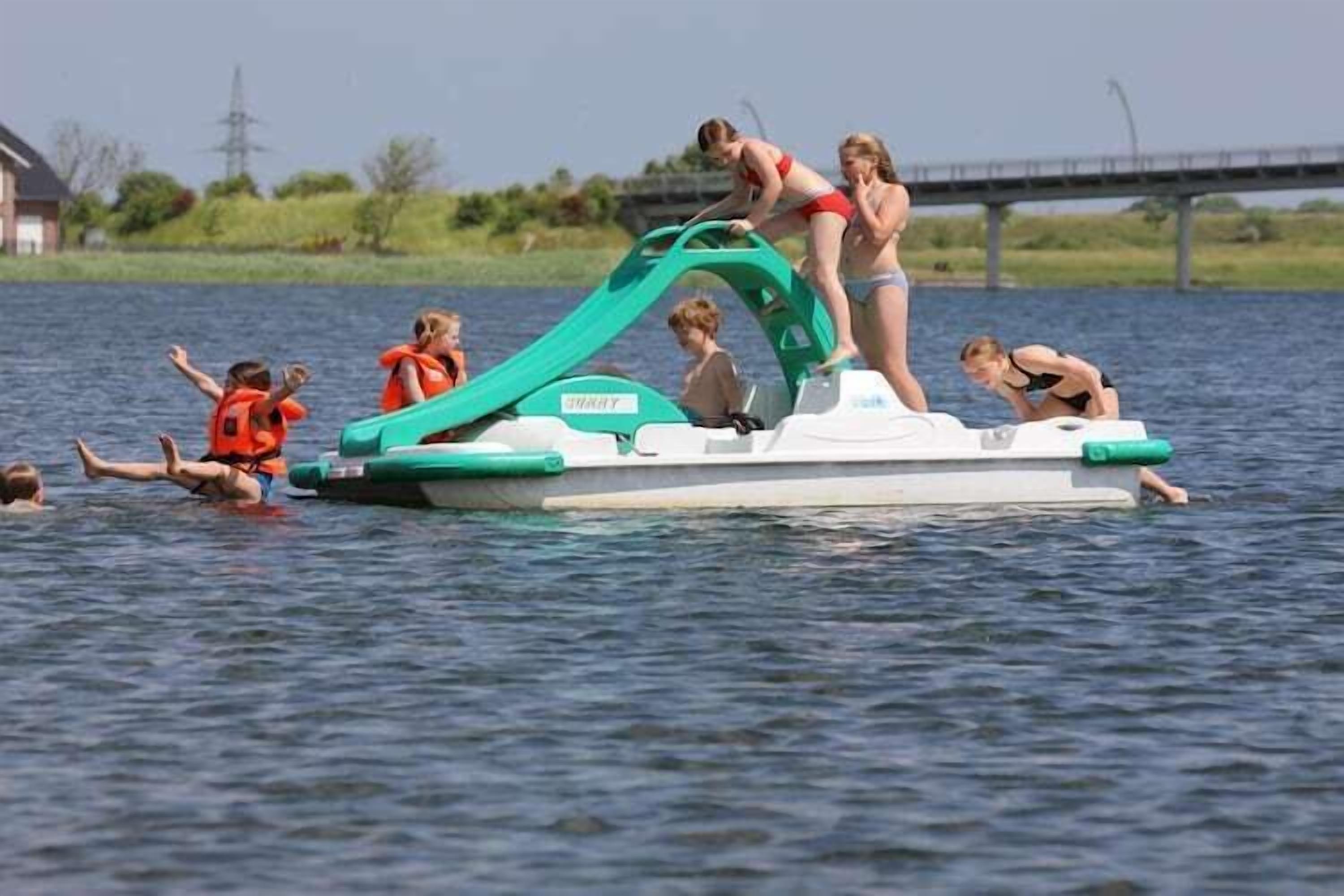 Eine Gruppe von Menschen in einem kleinen Boot mit einer grünen Rampe, einige tragen Rettungswesten, auf einem Gewässer mit einer Brücke im Hintergrund.