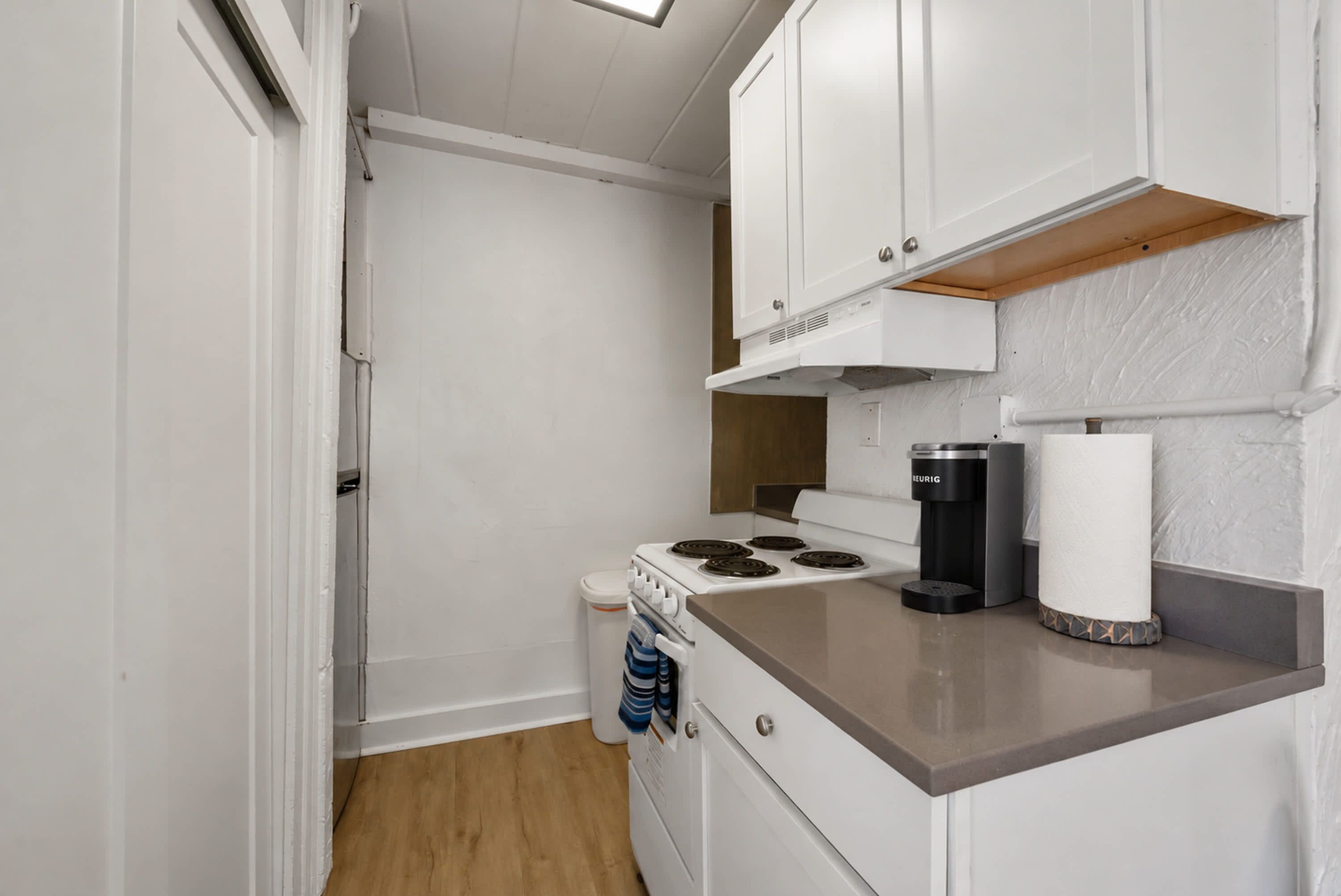 A compact kitchenette with white cabinets, a stove, a coffee maker, and a trash bin.