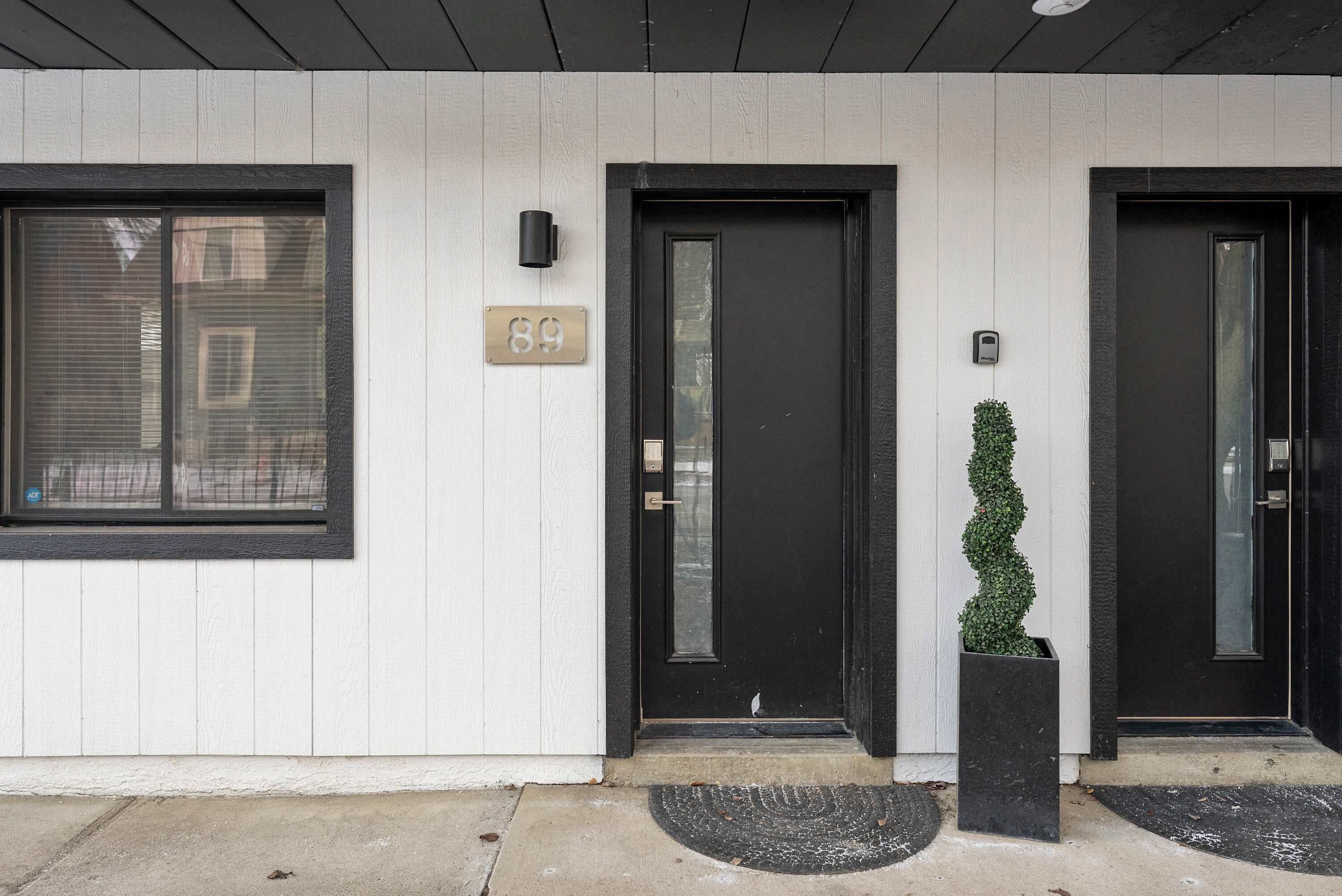 A building facade with a black door, a window with a number plaque, and a potted plant.