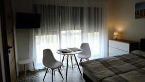 A hotel room with a bed, a desk with a lamp, a TV mounted on the wall, and a window with sheer curtains.