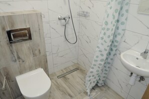 Un baño con inodoro, ducha con cabezal de ducha manual, lavamanos y pared con azulejos de mármol.