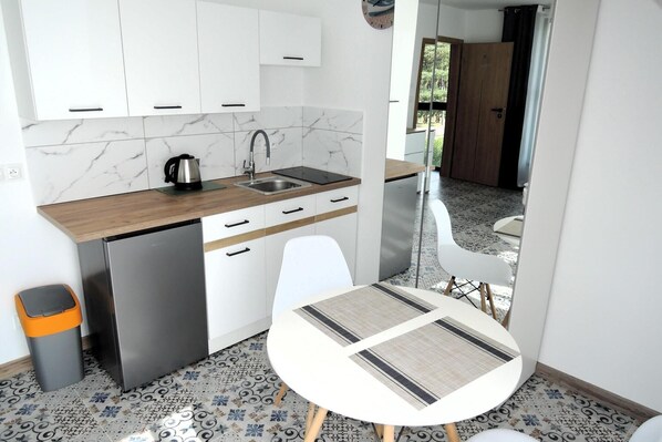 Eine kleine Küche mit weißen Schränken, einer Edelstahlspüle und einem kompakten Kühlschrank. Ein kleiner Tisch mit Flechtunterlage und ein weißer Stuhl sind vorhanden. Der Boden ist mit gemusterten Fliesen belegt, und ein Mülleimer ist sichtbar.