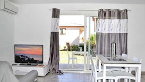 A modern living room with a TV, a dining table, and chairs, a sliding glass door leading to an outdoor area with a view of a building and greenery. - Rés. Marea Resort, Santa-Lucia-di-Moriani (Santa-Lucia-di-Moriani)