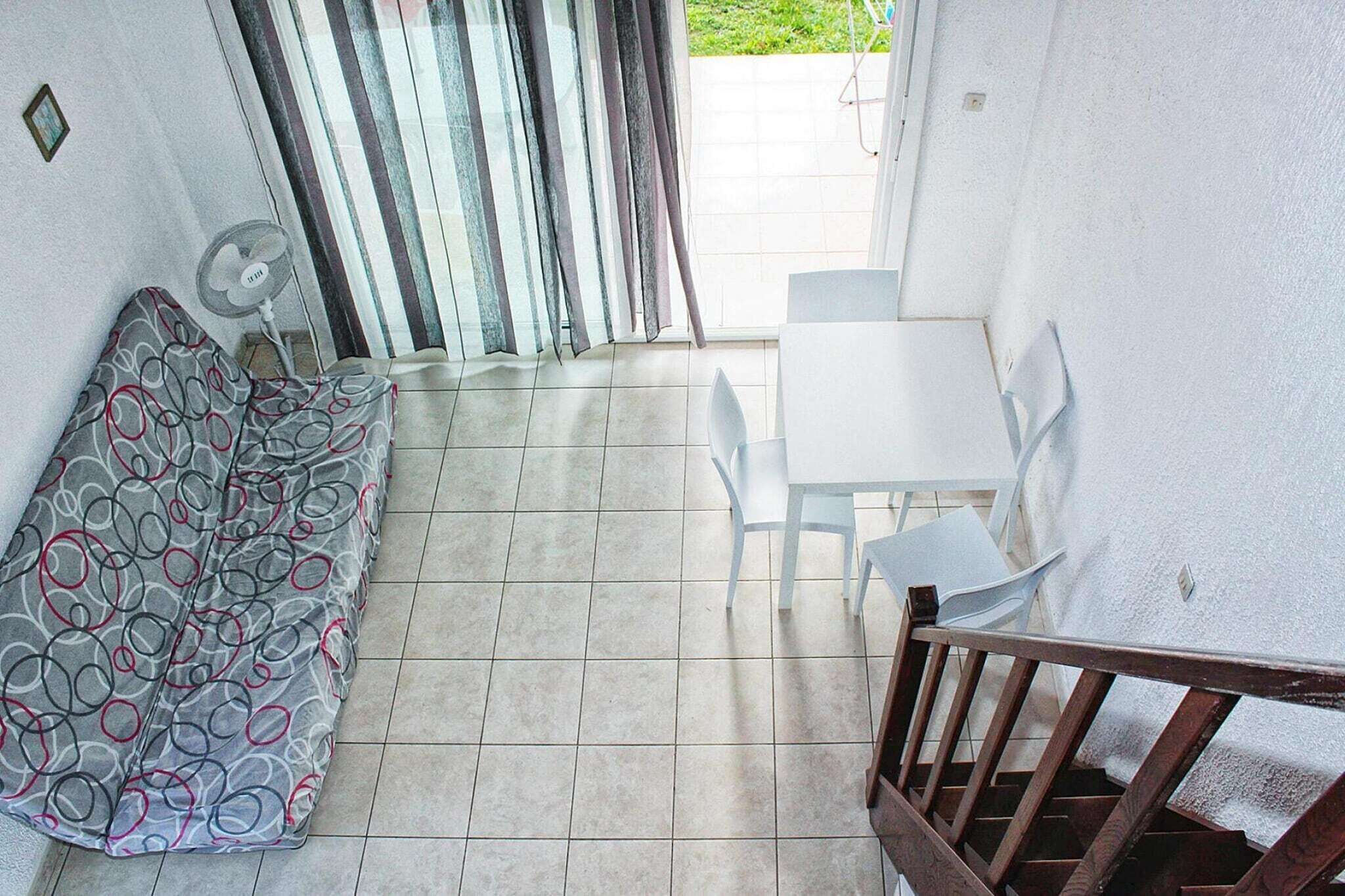 Ein kleiner Raum mit einer gemusterten Couch, einem weißen Tisch mit Stühlen, einem Ventilator und Blick auf einen Garten durch ein Fenster.