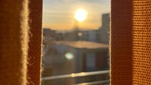 Une vue à travers une fenêtre dont le cadre est orné de motifs en relief, avec un paysage urbain et le soleil se couchant ou se levant en arrière-plan.