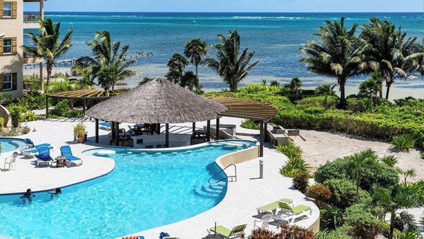 A resort pool with a thatched-roof gazebo, palm trees, and a view of the ocean. - Oceanview 2BR Condo- Pools, Beach, Spacious Balcony- Residences at Barrier Reef (San Pedro Town)