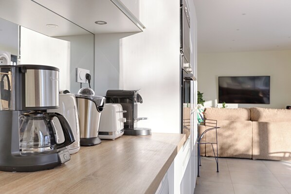 A modern kitchen with a coffee maker, toaster, and kettle. There is a wooden countertop and a view of a living area with a sofa and a TV. - Villa Carotte à 10 Minutes de Disneyland Paris (Chalifert)
